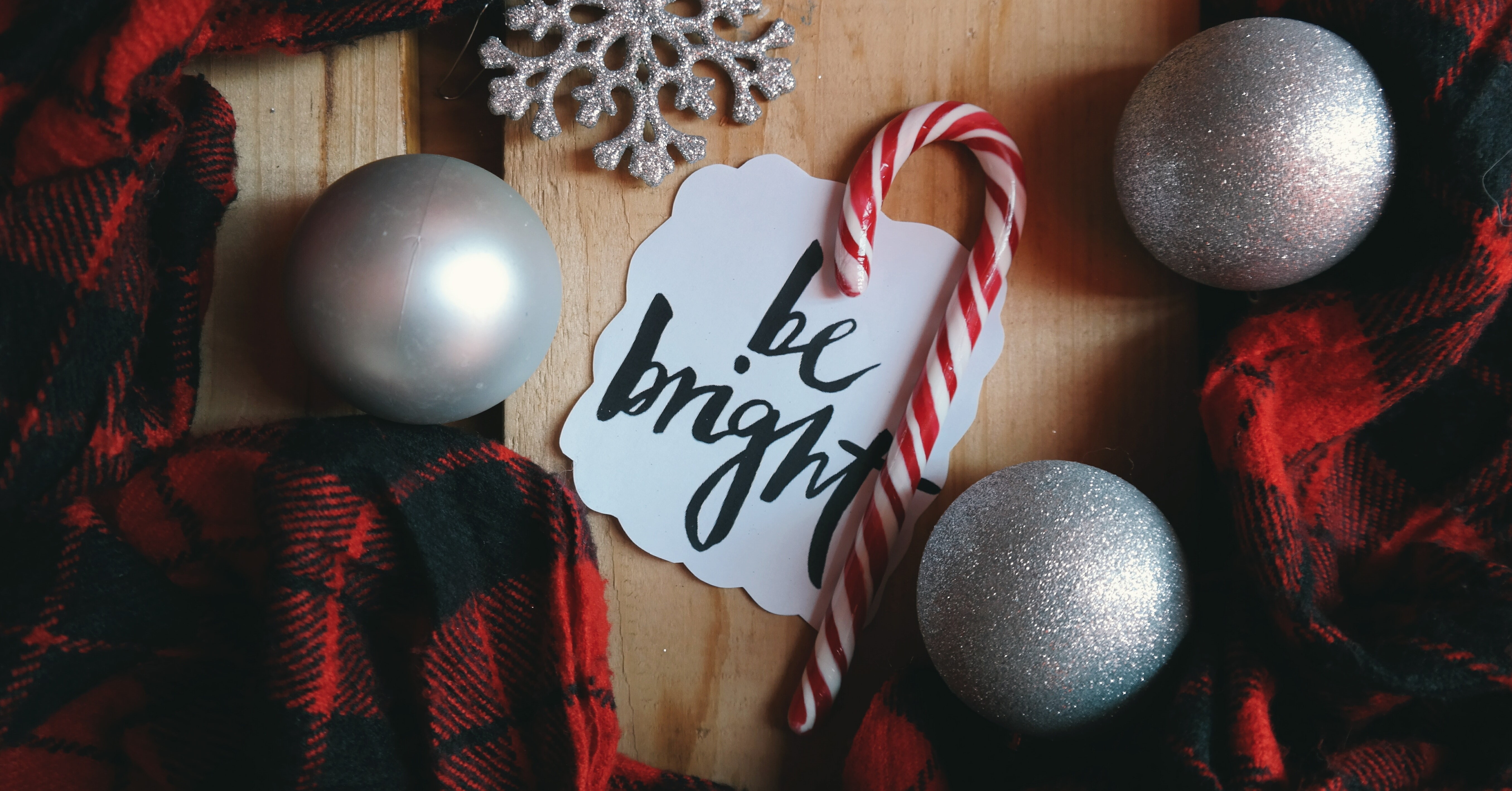 A candy cane on a wooden table. Table also has plaid cloth draped across with silver holiday ornaments. There is also a paper with calligraphy that reads "Be Bright."