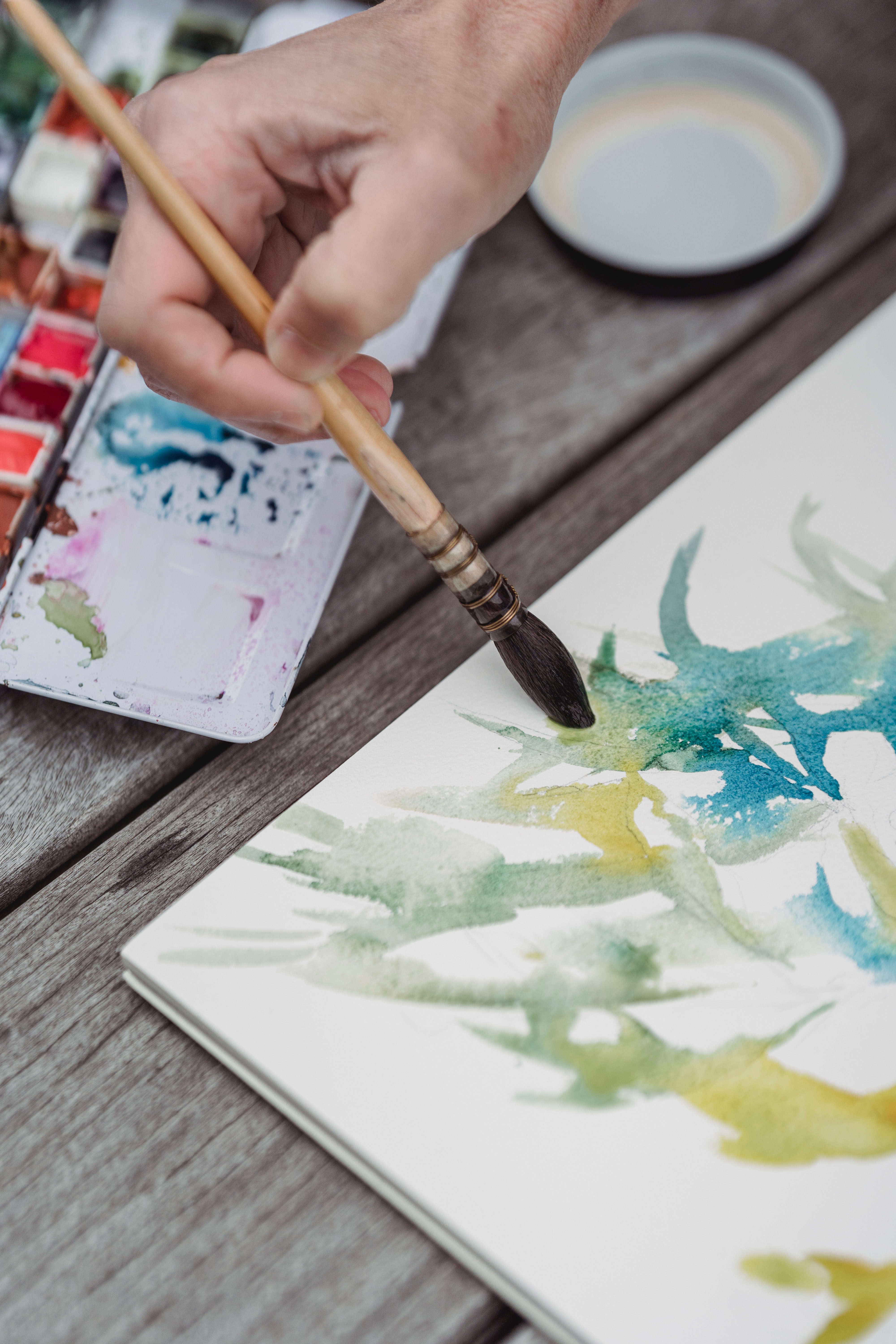 An artist holding a paintbrush on a canvas. There is blue, green, and yellow abstract shapes on the canvas and a watercolour palette in the background.