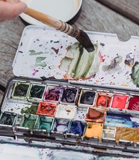 A messy watercolour paint palette with a paintbrush near it.